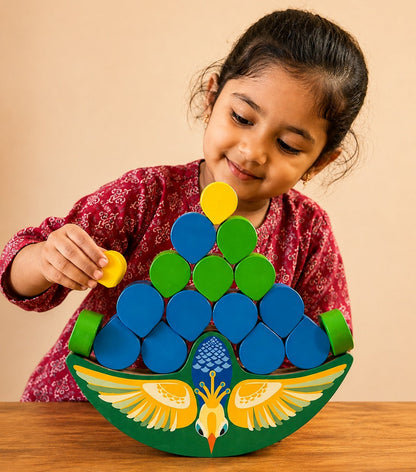 A young girl smiling while playing with a colorful wooden peacock-shaped stacking toy on a wooden table against a beige background.